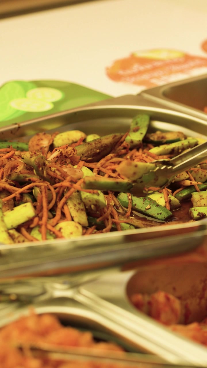 Una mano usa delle pinze di metallo per prendere kimchi di cetrioli fermentati da un vassoio di un buffet di ristorante.