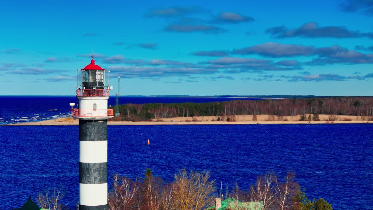 A tall striped lighthouse stands against brilliant blue Baltic Sea and sandy shore, surrounded by bare trees and a bright sky, symbolizing navigation and coastal heritage.