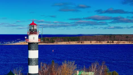 a tall striped lighthouse stands against brilliant blue baltic sea and sandy shore, surrounded by bare trees and a bright sky, symbolizing navigation and coastal heritage.