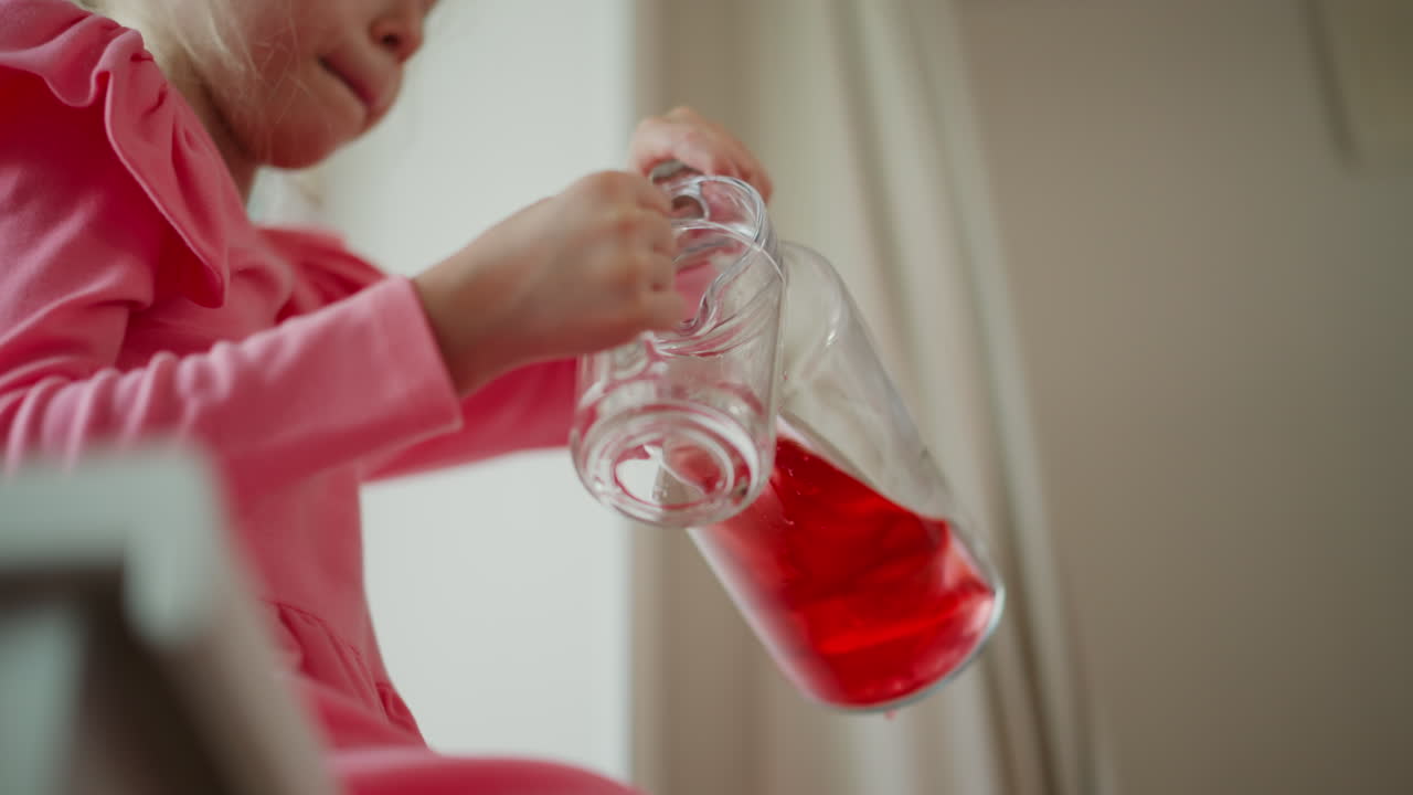 Low angle of petite figure seated by window in kitchen corner drinking red juice from clear cup, small hands holding mug, soft pink dress, wooden drawer in background and gentle daylight glow