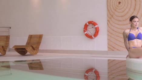 woman sits leisurely on edge of indoor pool at wellness centre, panning shot