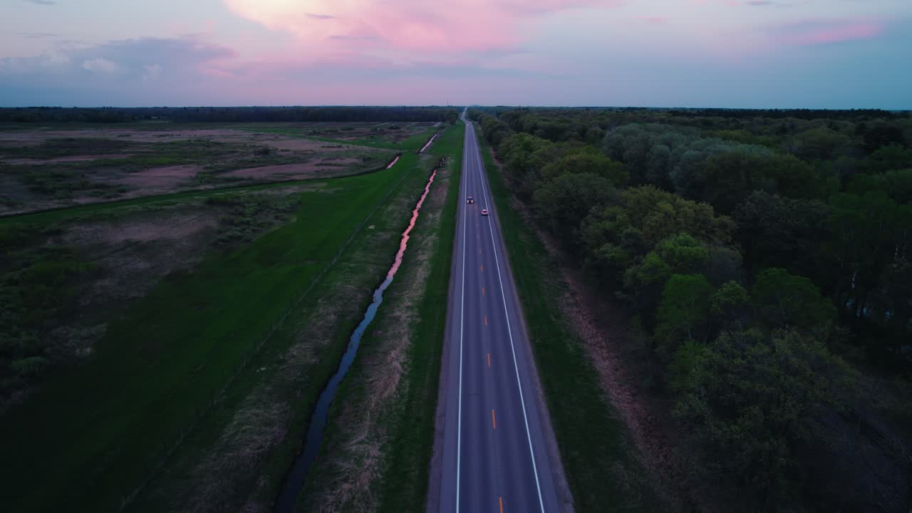 Drone tracks a car in reverse over a quiet North Wisconsin road at sunset, with pink skies, green fields, and tranquil rural scenery.