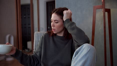 a tired brunette girl in a gray jacket drinks tea from a cup and thinks about life while sitting in a cozy room in a modern apartment