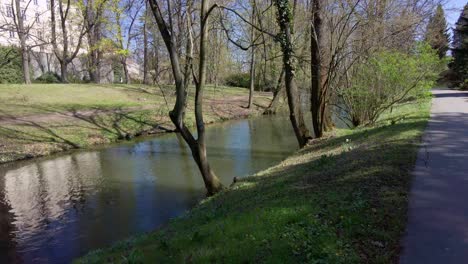 Río-Que-Fluye-Con-Bancos-Cubiertos-De-Hierba-Y-árboles,-En-El-Fondo-La-Gente-Pasa-Por-Las-Murallas-Y-Edificios-Históricos,-Olomouc