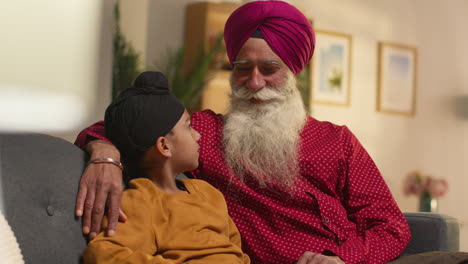 un abuelo y un nieto sikh sonrientes con turbantes sentados en el sofá en casa hablando juntos 1