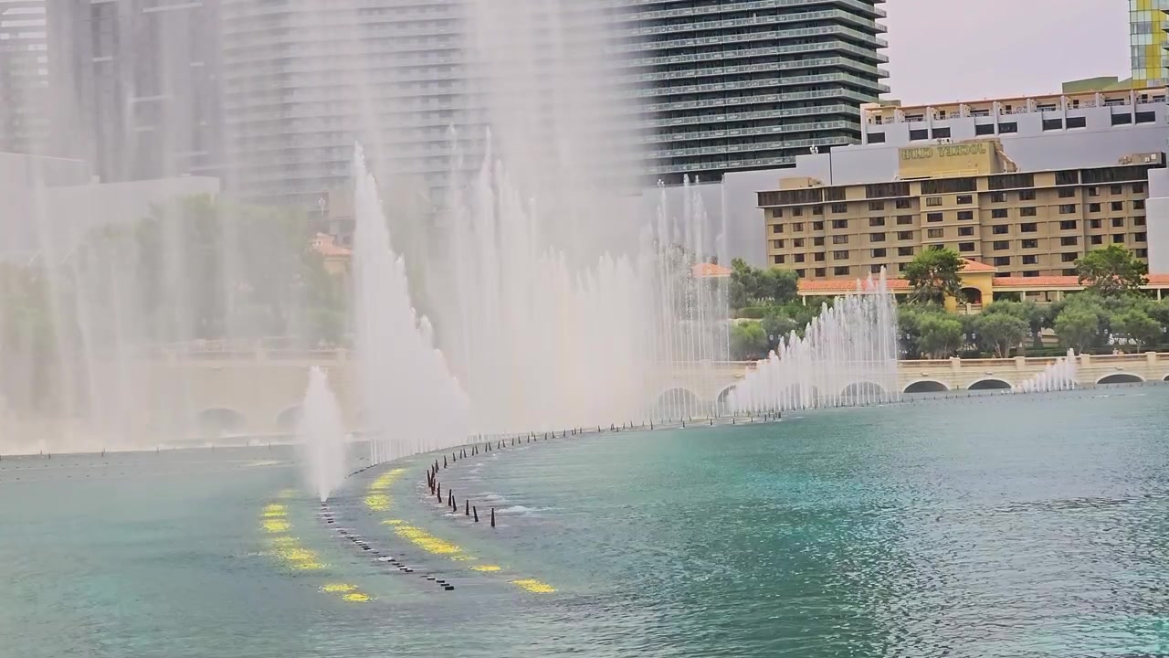 Vista panorámica de un lago con fuente, con altos chorros de agua, reflejos azul verdosos, arcos de piedra, árboles paisajísticos, torres de hotel y un ambiente diurno luminoso en un resort de lujo para videos de viajes