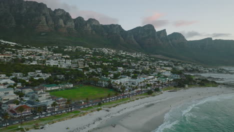 Forwards-fly-above-sea-coast,-waves-washing-sand-beach.-Buildings-in-tropical-vacation-destination,-mountains-in-background.-Cape-Town,-South-Africa
