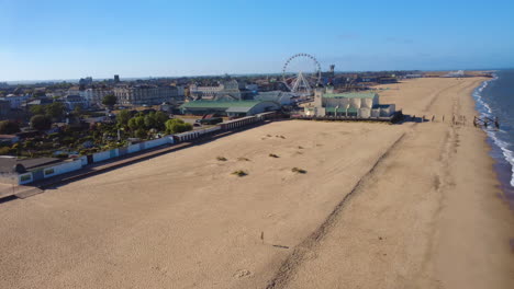 aerial drone footage circling wellington pier in great yarmouth, norfolk coast. it captures a classic british seaside with sandy beach, amusements, arcades, and offshore wind turbines on the horizon