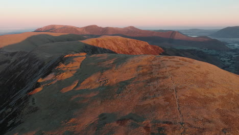 Flying-over-mountainside-lit-by-autumn-dawn-sunshine-revealing-wider-mountainous-landscape-beyond