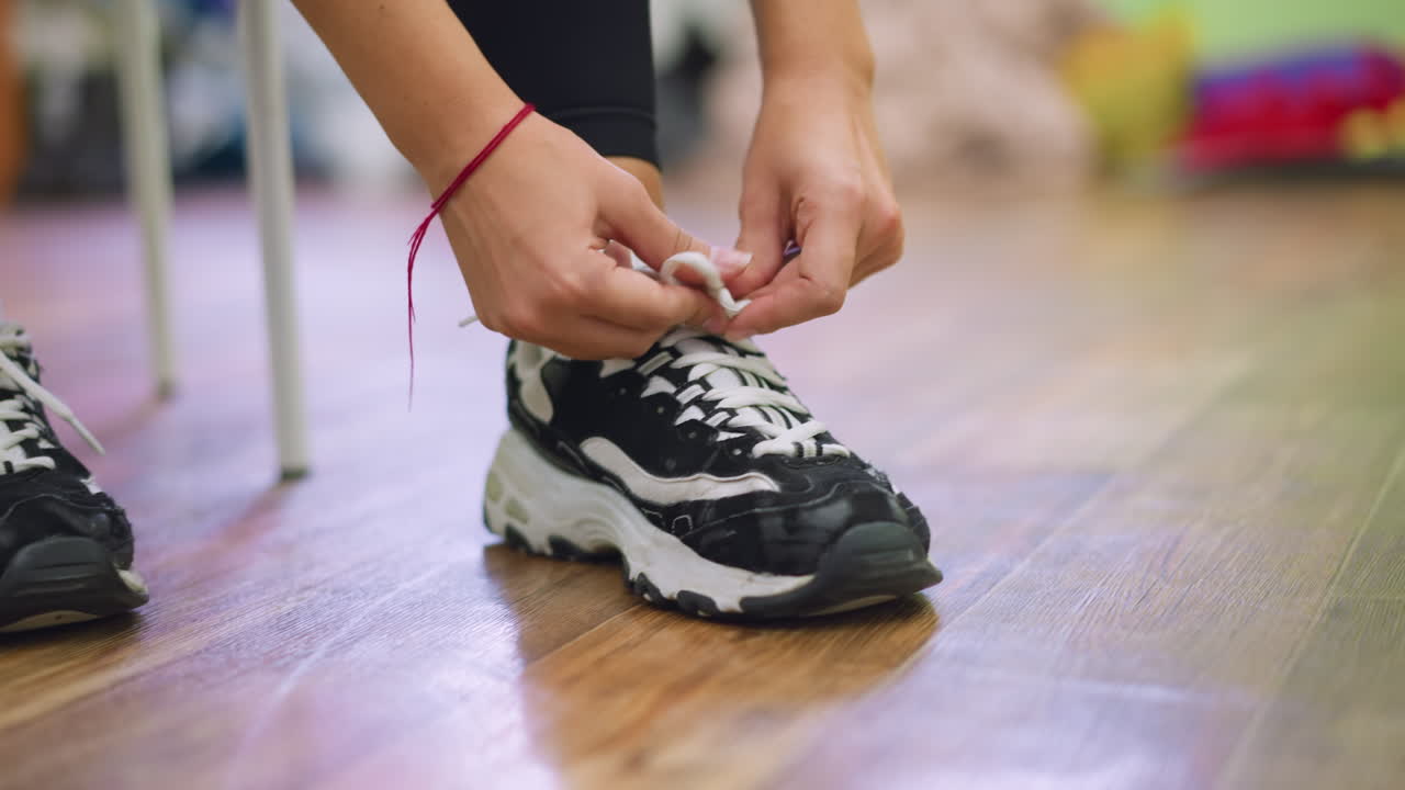 Close view of woman in black sneakers tying laces on wooden floor, hand wearing red string bracelet, detail capturing moment of preparation for training with focus on footwear and movement indoors