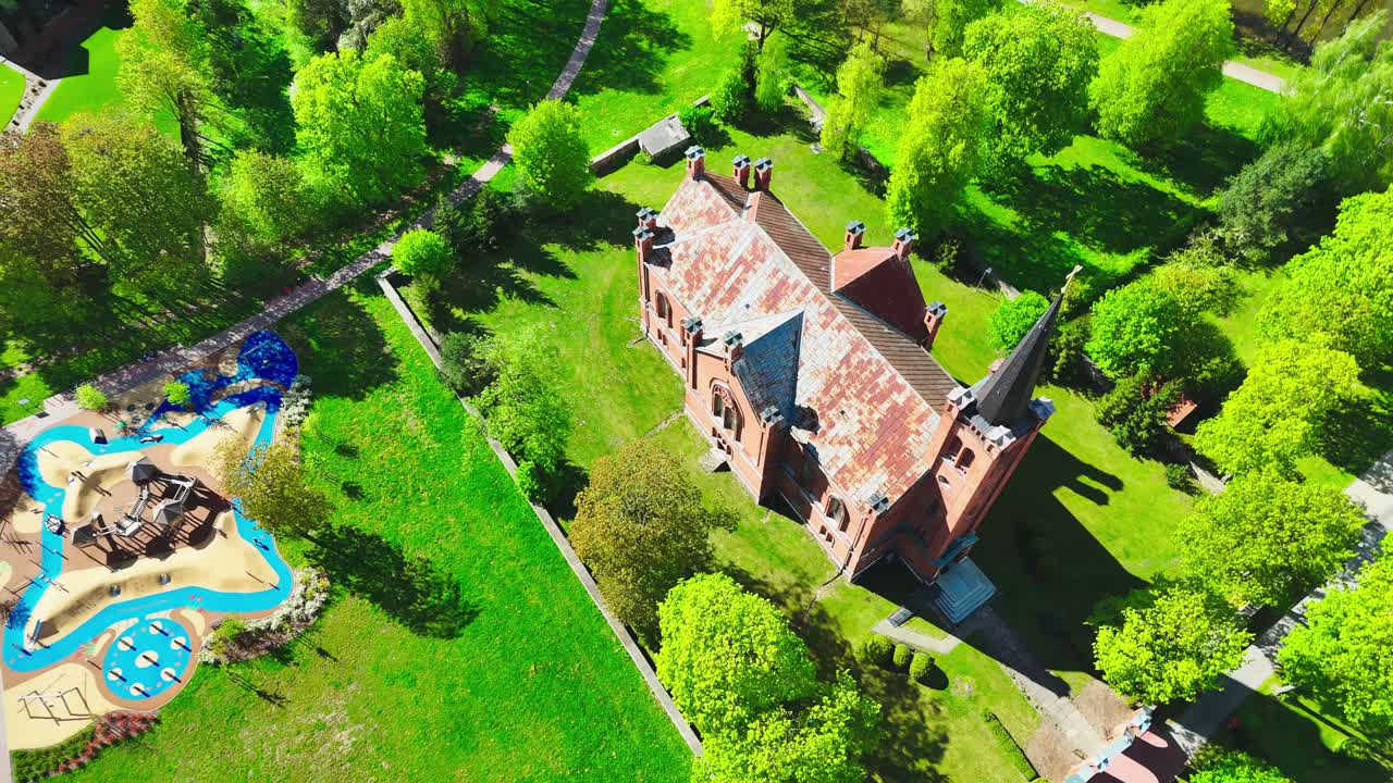 A red brick gothic-style manor with pointed turrets sits in a lush green park beside a vibrant children's playground, surrounded by spring trees and walking paths.