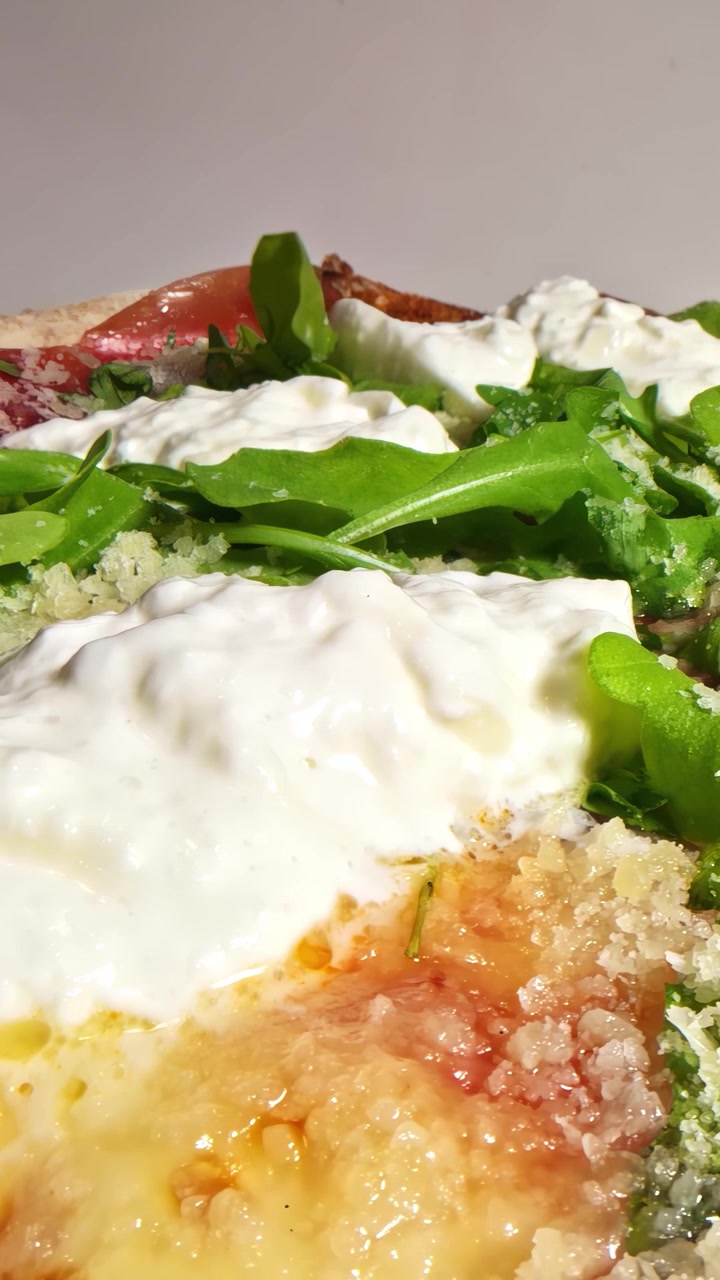 Un primo piano di una pizza appena sfornata guarnita con cremosa burrata, foglie fresche di rucola, sottili fette di prosciutto crudo e Parmigiano grattugiato su una crosta croccante. Video verticale, Social media
