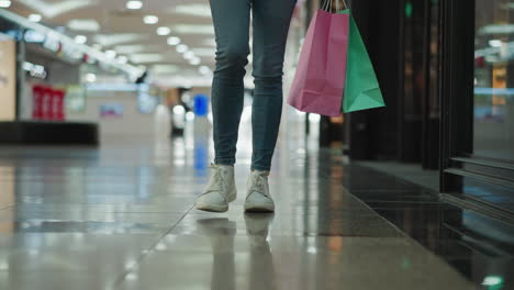 leg view of a woman in jeans and canvas sneakers carrying mint and pink shopping bags in her right hand while walking through a brightly lit shopping mall with glossy tiled floors