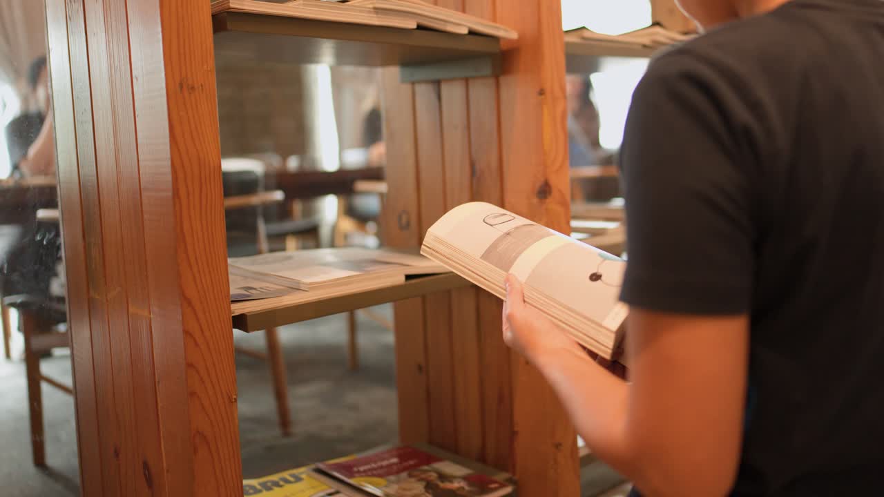 En person står ved en træbogreol og bladrer i et magasin på en lys café.