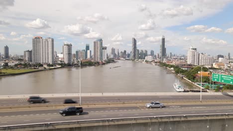 Bangkok-Skyline-Along-Chao-Phraya-River,-Aerial-view-of-Traffic-on-Bridge-Road,-Thailand