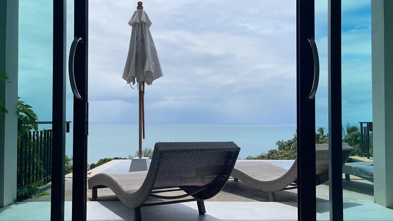 Relaxing oceanfront patio with curved lounge chairs and closed umbrella facing sea, framed by open glass doors and lush greenery under clear sky in Ko Samui