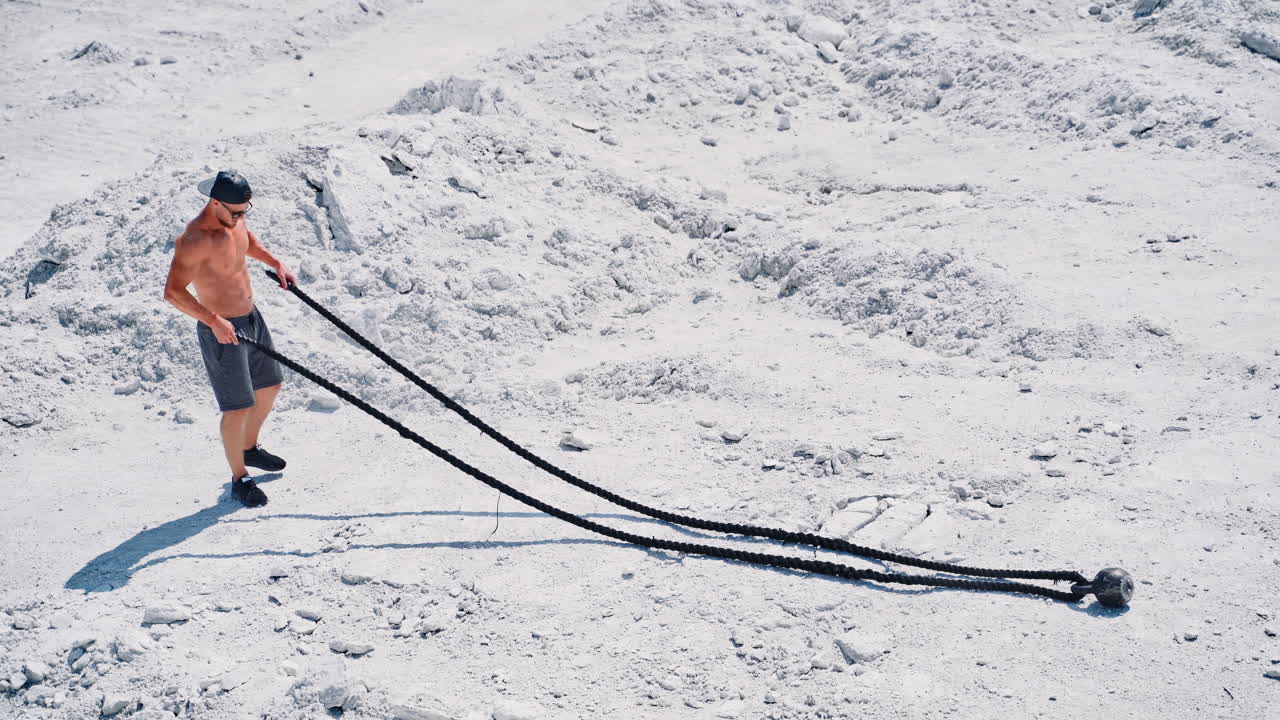 fitness athlete in a cap and sunglasses prepares for training with ropes outdoor. Heavy training in a quarry. Intense workout with ropes in white mountains. Strong attractive bodybuilder. Lifestyle. White landscape.