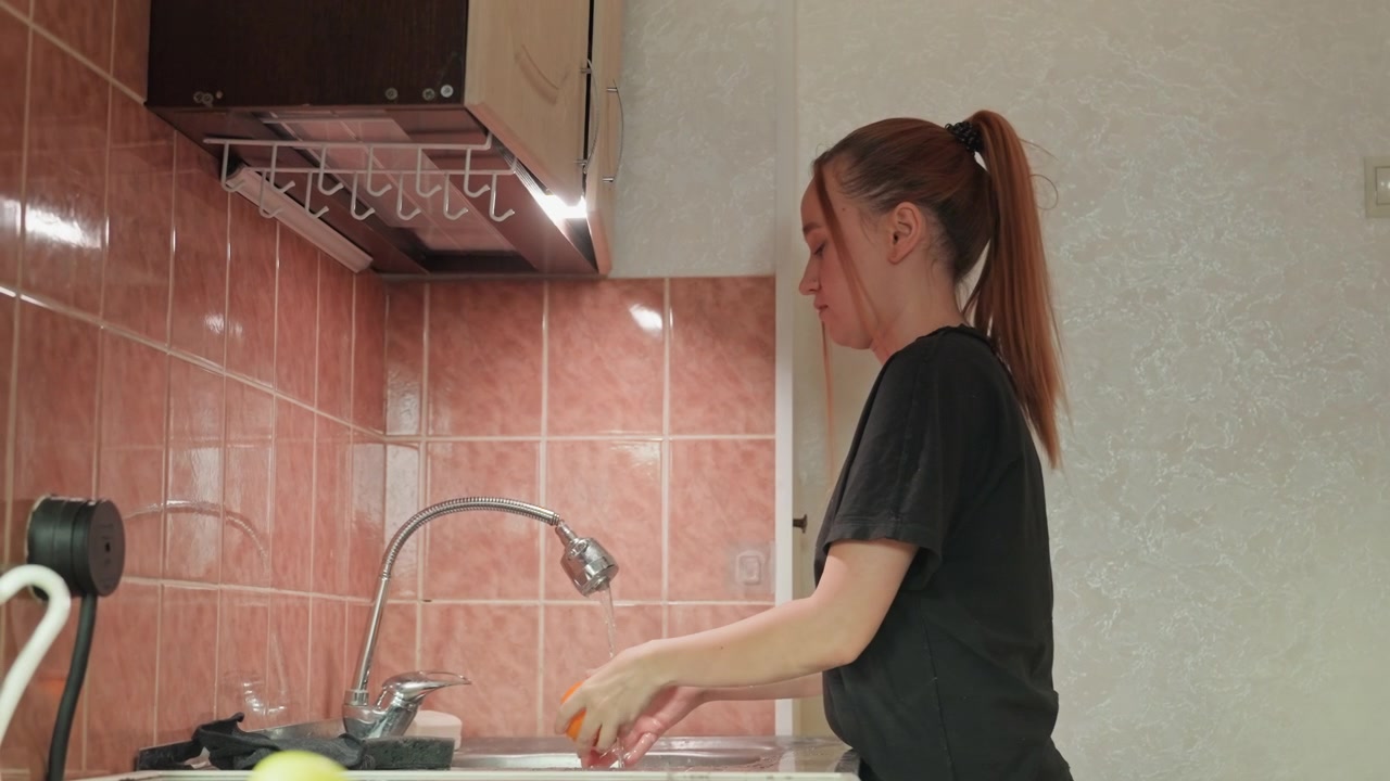 Una donna si posiziona rivolta a sinistra mentre tiene un oggetto. Vari accessori da cucina come una tazza, apparecchi elettrici neri. Ambiente cucina con pareti piastrellate rosa e colori chiari.