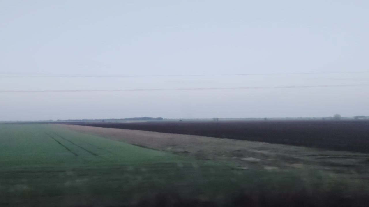 A professional tracking shot captures the expansive, dark fields of the Alföld under a pale morning sky. The rhythmic horizon passes by during an early journey from Keleti toward Gyomaendrőd.
