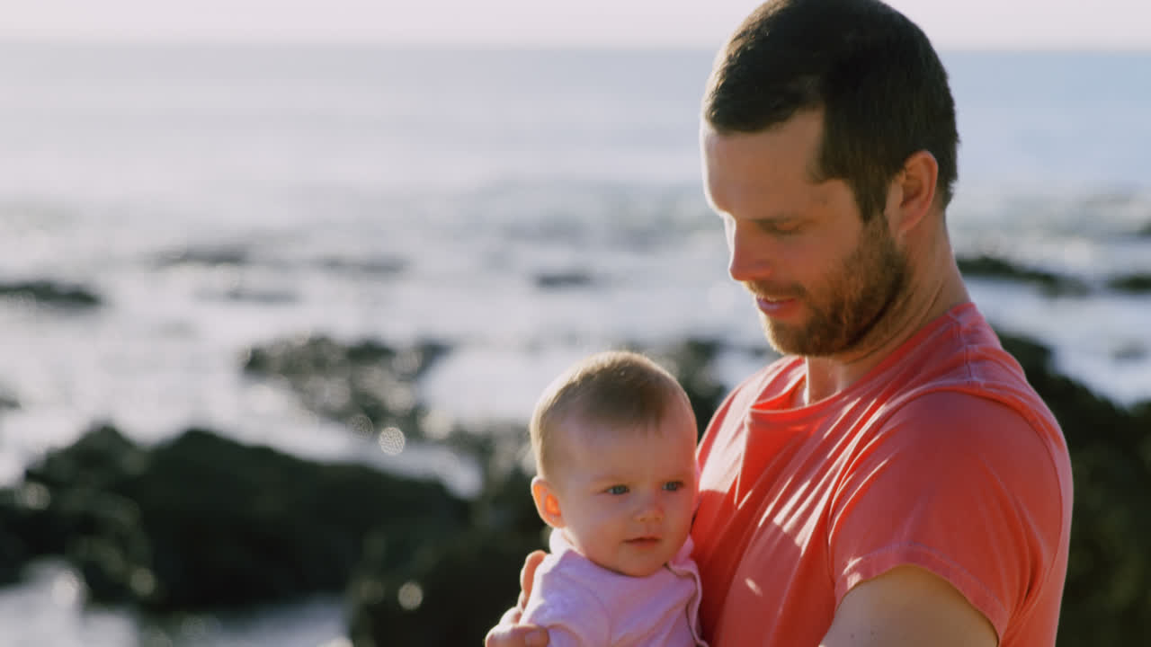 Seitenansicht eines kaukasischen Vaters mittleren Alters, der an einem sonnigen Tag ein Baby am Strand hält 4K