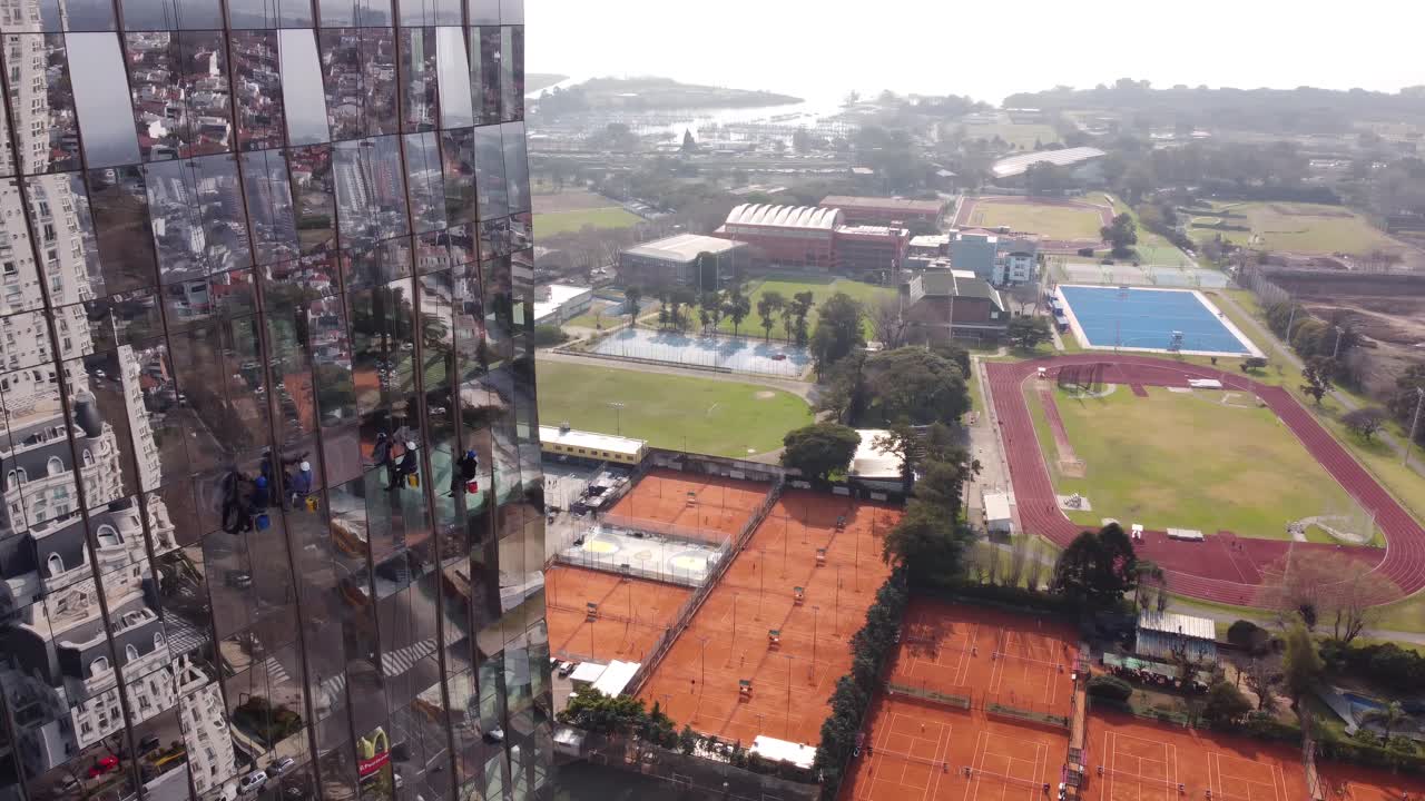 tiro de dron de lavadora de ventanas limpiando ventanas espejadas de rascacielos durante el sol - en canchas de tenis de fondo y campo de fútbol en buenos aires, argentina