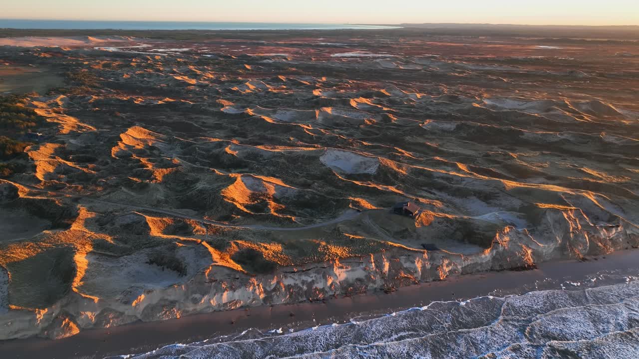 vista aérea de grandes dunas de arena de pie junto al mar iluminadas por la luz del atardecer, una casa de verano se encuentra en las dunas, y el agua del mar lava la costa