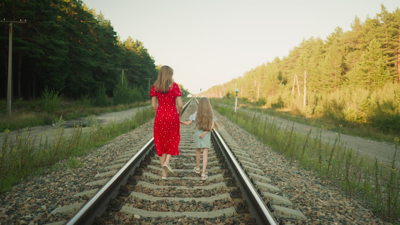rear view of woman in red dress holding young girl hand as they walk along railway track surrounded by forest trees and rural paths during soft daylight, child placing other hand behind back