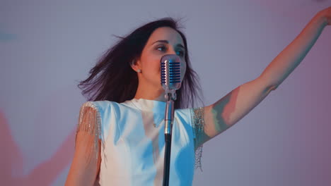 singer in a white gown smiling with hands up in the air, body moving expressively while performing into a vintage microphone. set against a background of pink and blue