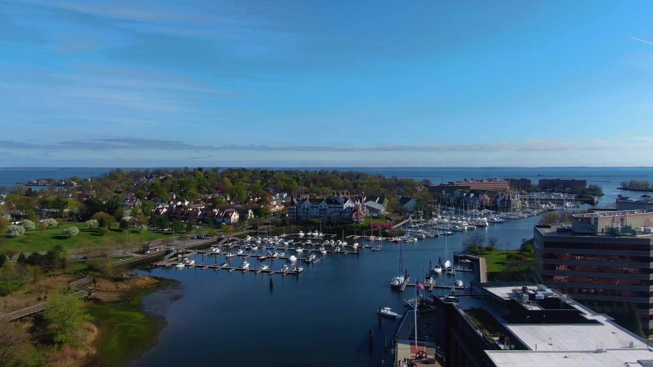 Toma de dron moviéndose hacia la izquierda a través de un puerto deportivo y un vecindario frente al mar con barcos, casas y árboles durante el día de primavera en Stamford, Connecticut
