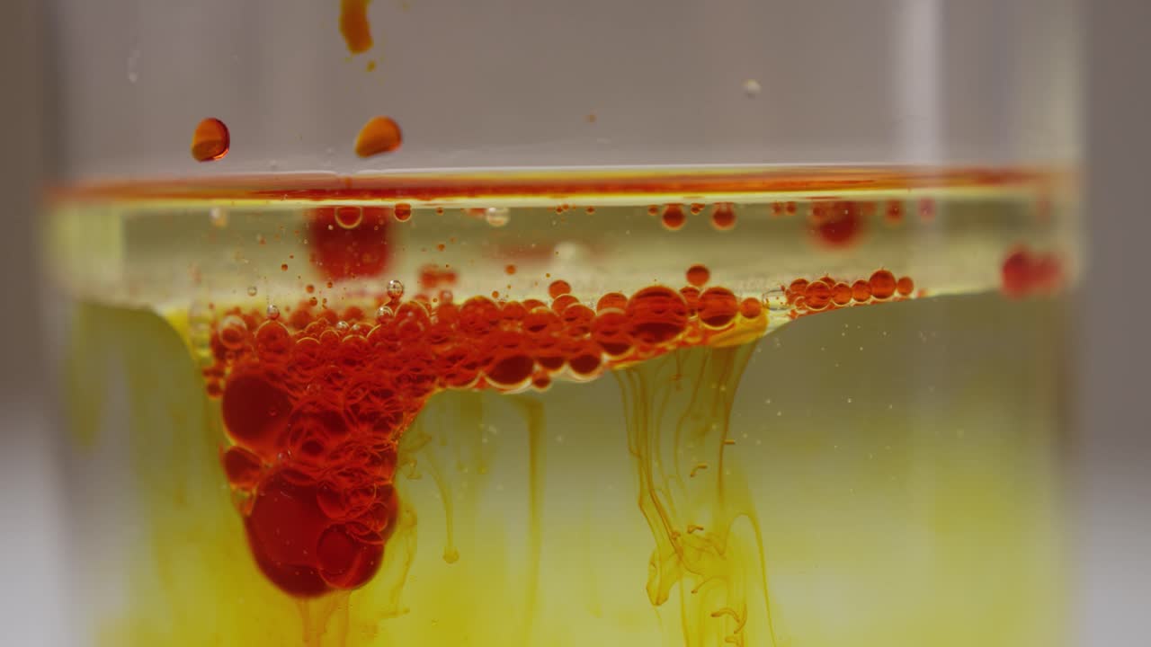 Slow motion shot of an oil emulsion swimming on the surface of a water filled glass container that is infested by another red liquid that slowly sinks below the oil layer