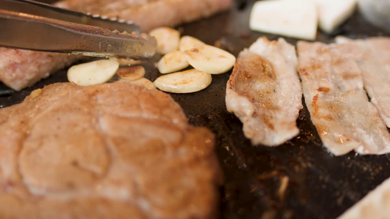 Tongs pick sliced garlic beside pork belly on hot grill, shallow focus, warm indoor lighting