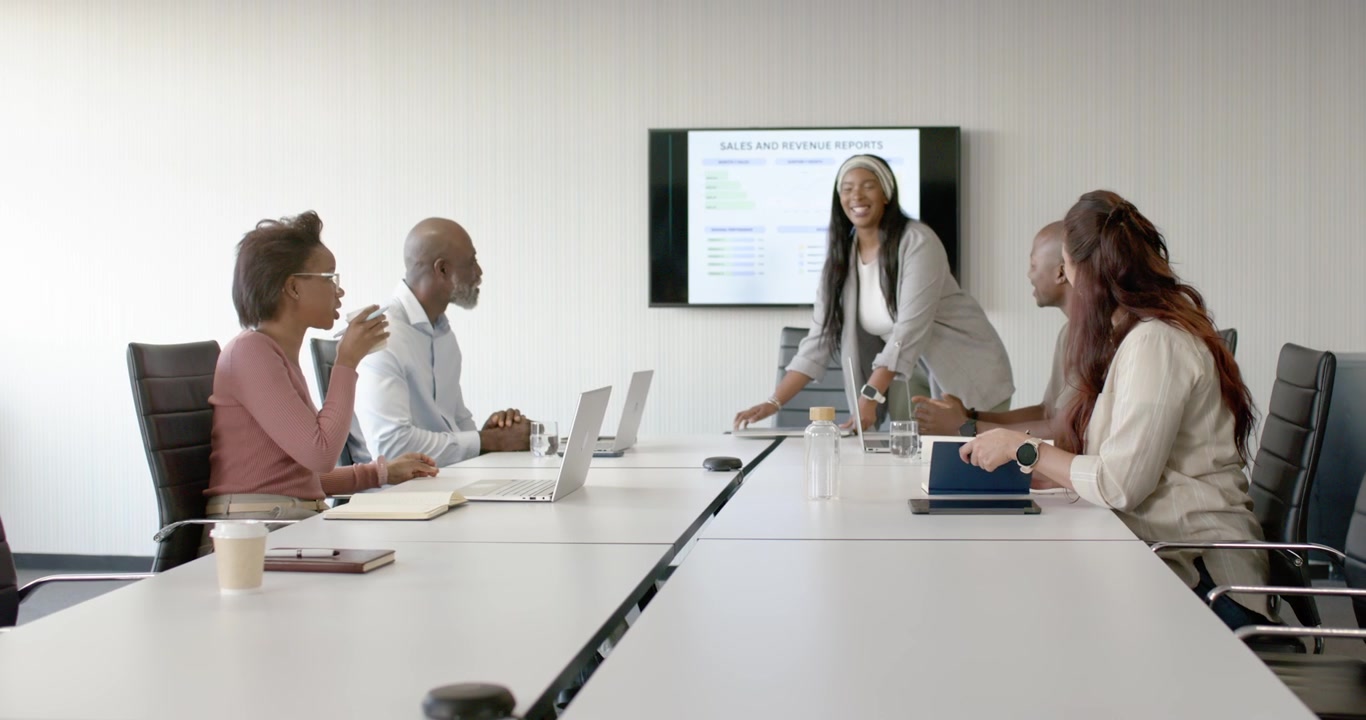 Diverse team watching African American presenter walking to front at meeting table presenting sales