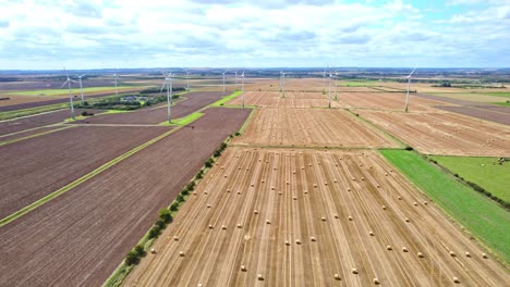 Oben-Offenbart-Ein-Luftvideo-Einen-Faszinierenden-Anblick:-Windkraftanlagen-Drehen-Sich-Anmutig-Auf-Dem-Kürzlich-Abgeernteten-Feld-Eines-Bauern-Aus-Lincolnshire,-Im-Vordergrund-Goldene-Heuballen