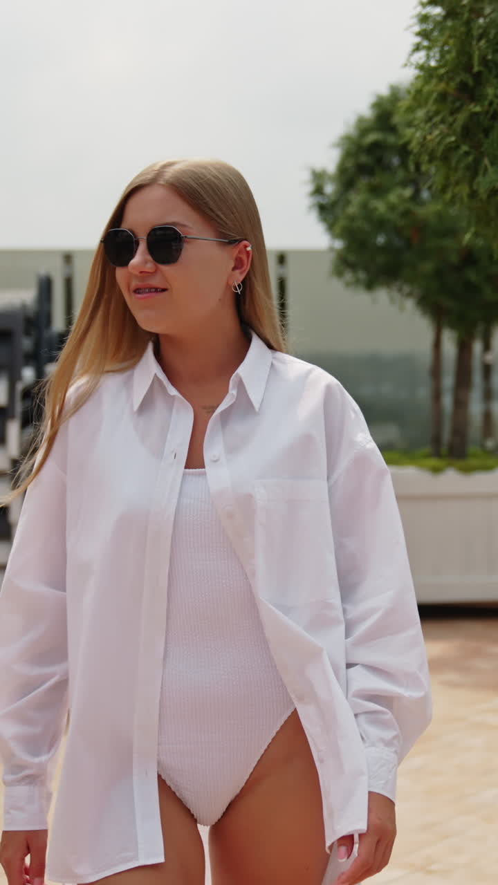 Beautiful long-haired young lady wearing white swimsuit and shirt walks slowly outdoors. Blonde woman wearing sunglasses smiles to the camera. Vertical video