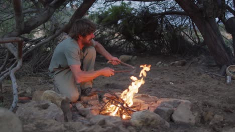 feliz barbudo caucásico hombre sobreviviente añadiendo leña al fuego en el campamento en el desierto