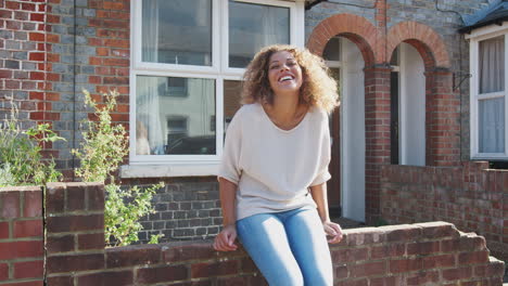 Portrait-Of-Young-Woman-Sitting-On-Wall-Outside-New-Home-In-Urban-Street