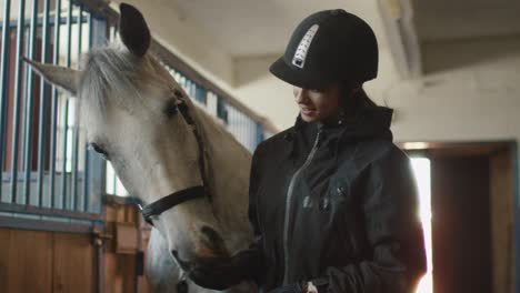 young jockey girl is stroking a white horse in a stable.