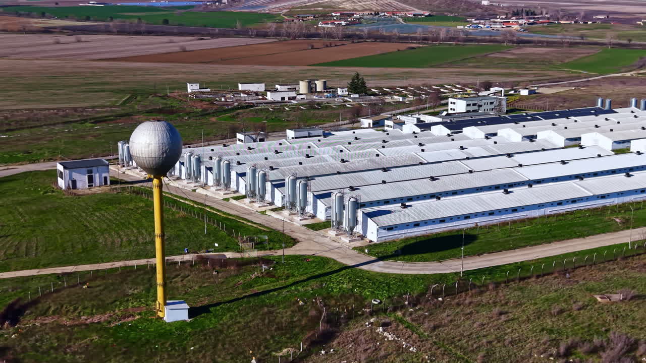 Grande impianto industriale situato in un'area rurale con campi verdi e cielo sereno, dove i lavoratori svolgono compiti e operazioni quotidiane per esigenze di produzione e logistica