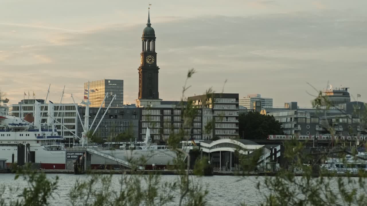 toma de grúa que revela una vista de la torre de la iglesia de san miguel, hamburgo, cruzando el río elba al atardecer contra un cielo nublado visto a través de una frondosa vegetación.