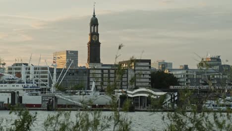 crane shot revealing a view of the spire of st michaels church, hamburg across the river elbe at sunset against a cloudy sky viewed through leafy greenery