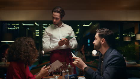Handsome-waiter-helping-couple-in-restaurant.-Multiethnic-lovers-looking-menu.
