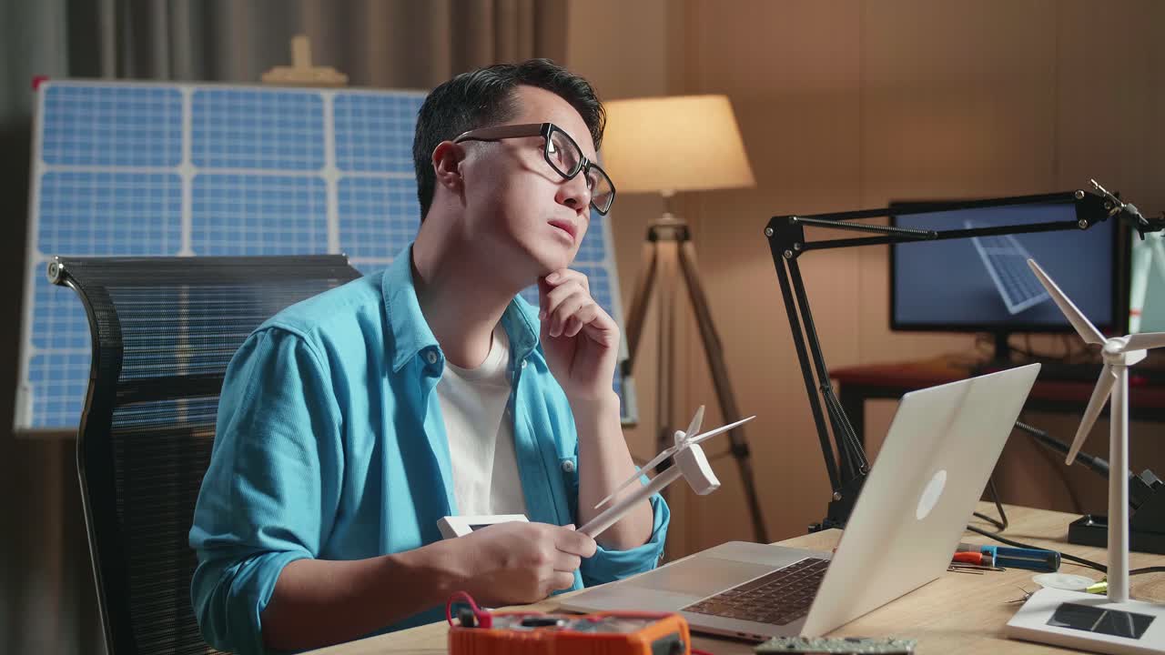 アジア人男性が太陽電池の前に座って風力タービンを見てオフィスでラップトップで作業している間考えています