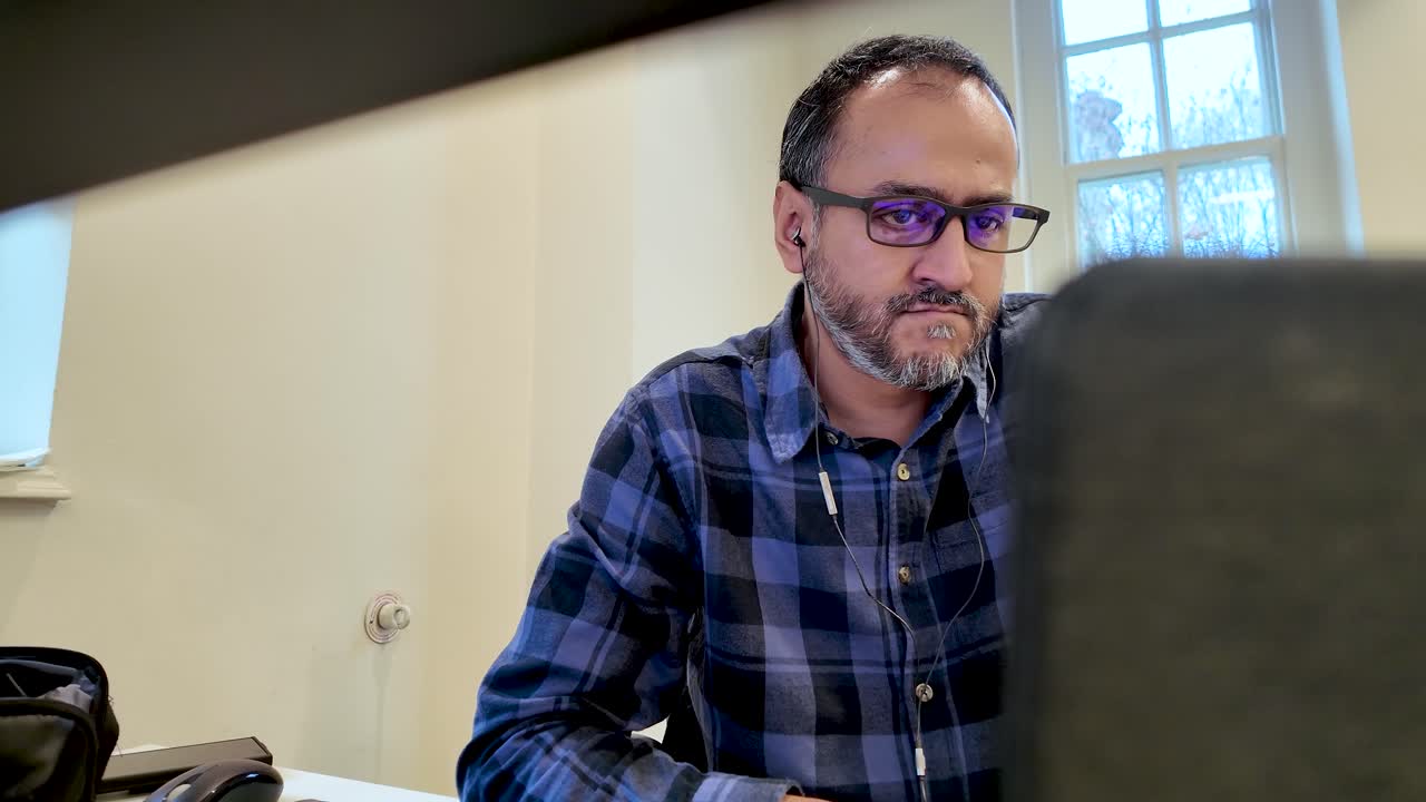 Südasiatischer Mann mit Brille und Kopfhörern, der sich auf seinen Laptop konzentriert, während er arbeitet