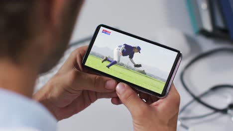 compuesto de un médico masculino viendo un juego de béisbol en un teléfono inteligente