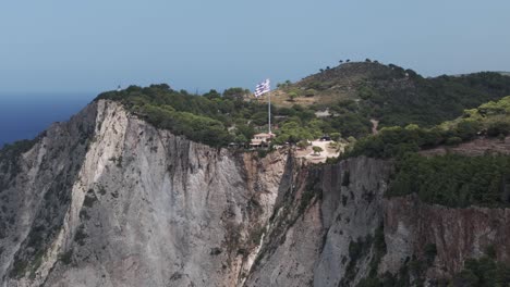 Vista-Aérea-De-La-Bandera-Nacional-Griega-Ondeando-En-La-Cima-De-Una-Colina-Sobre-Los-Acantilados-Y-El-Mar-Jónico,-Isla-De-Zakynthos-50fps