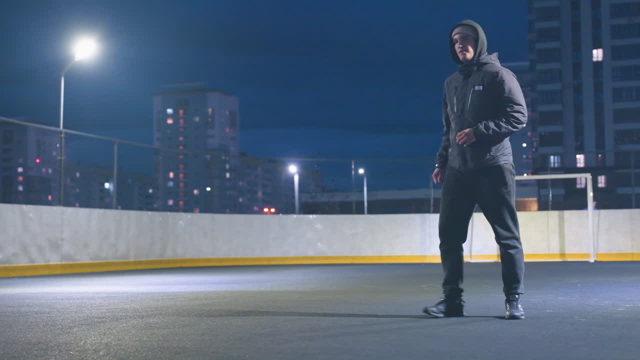 Man in hoodie kicks soccer ball on illuminated field under street lights with goal post in background, cityscape featuring tall buildings creates dynamic urban ambiance during nighttime
