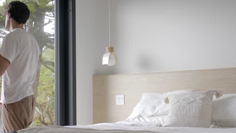 man sitting on bed stands up and opens curtains, allowing morning sunlight through glass window into the bedroom at the hotel