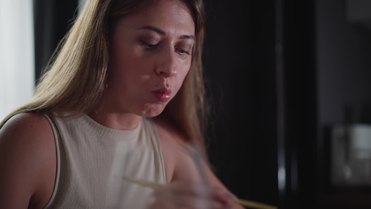 Hungry woman in sleeveless gown eats cake using chopsticks, eyes partially closed as she takes a big bite, indoor setting with soft lighting and blurred kitchen background
