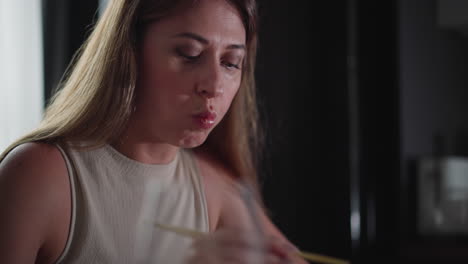 hungry woman in sleeveless gown eats cake using chopsticks, eyes partially closed as she takes a big bite, indoor setting with soft lighting and blurred kitchen background