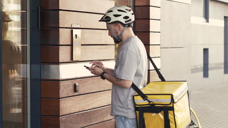 food delivery guy wearing thermal backpack calls the entry phone of a building to make a delivery